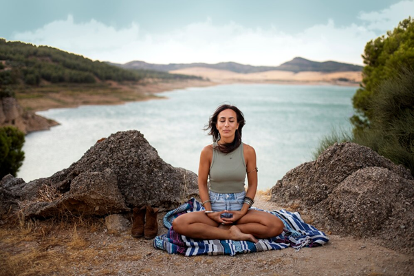 Long brown haired lady sat cross legged on a blue stripy blanket on a beach with a bay in the background.