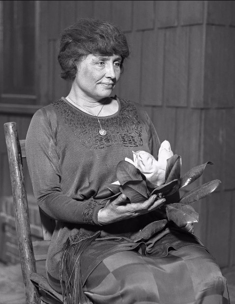 Helen_Keller_circa_1920 Helen_Keller_circa_1920 sitting in a chair looking forward with a large flower in her lap.