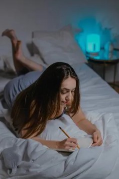 Bedtime_Writing_Laid_Down Woman with long brown hair journaling while laid on her front on a bed.