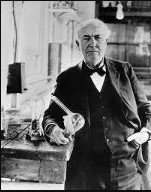 Thomas_Edison-0419860 Black and white photo of Thomas Edison holding some plans with a lightbulb hanging behind him.