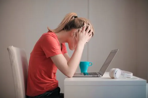 Lady with blonde hair in a ponytail sitting at a desk with her head in her hands looking at a laptop.
