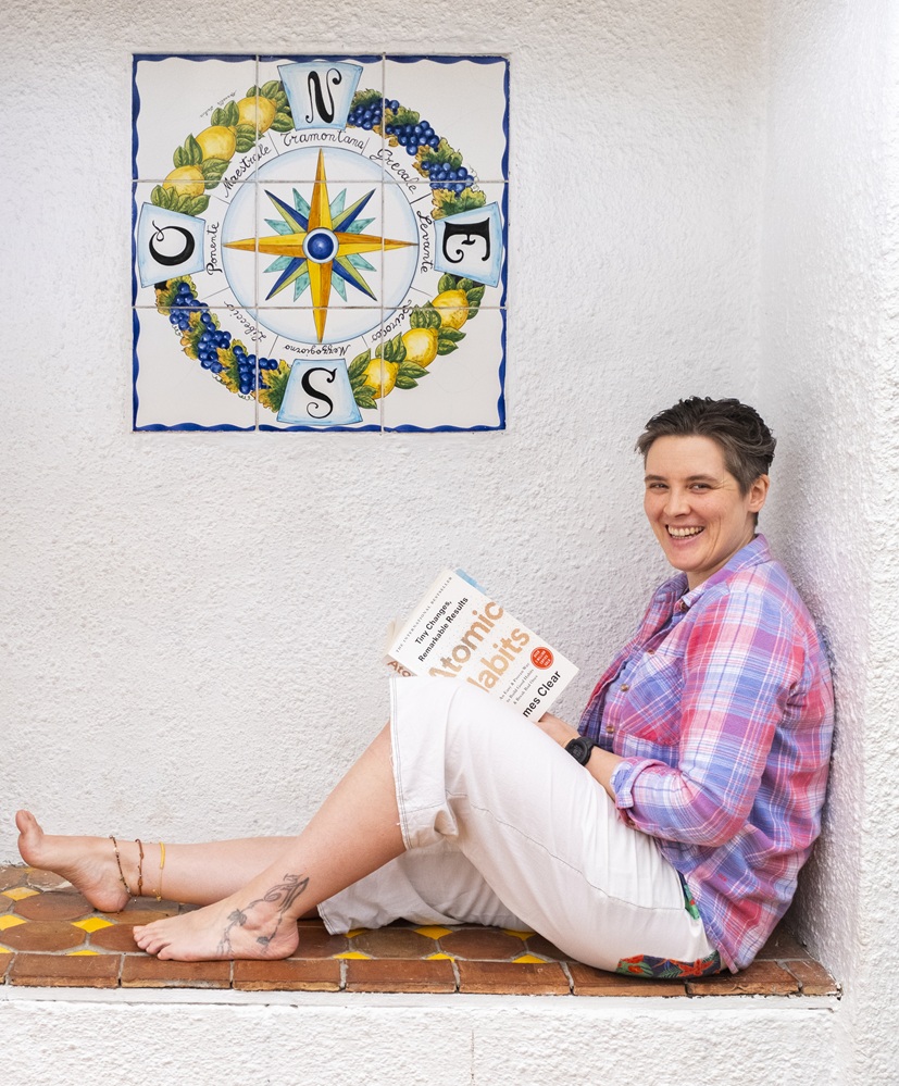 Jaiye sat in a white walled area with a compass rose on the wall reading Atomic Habits.
