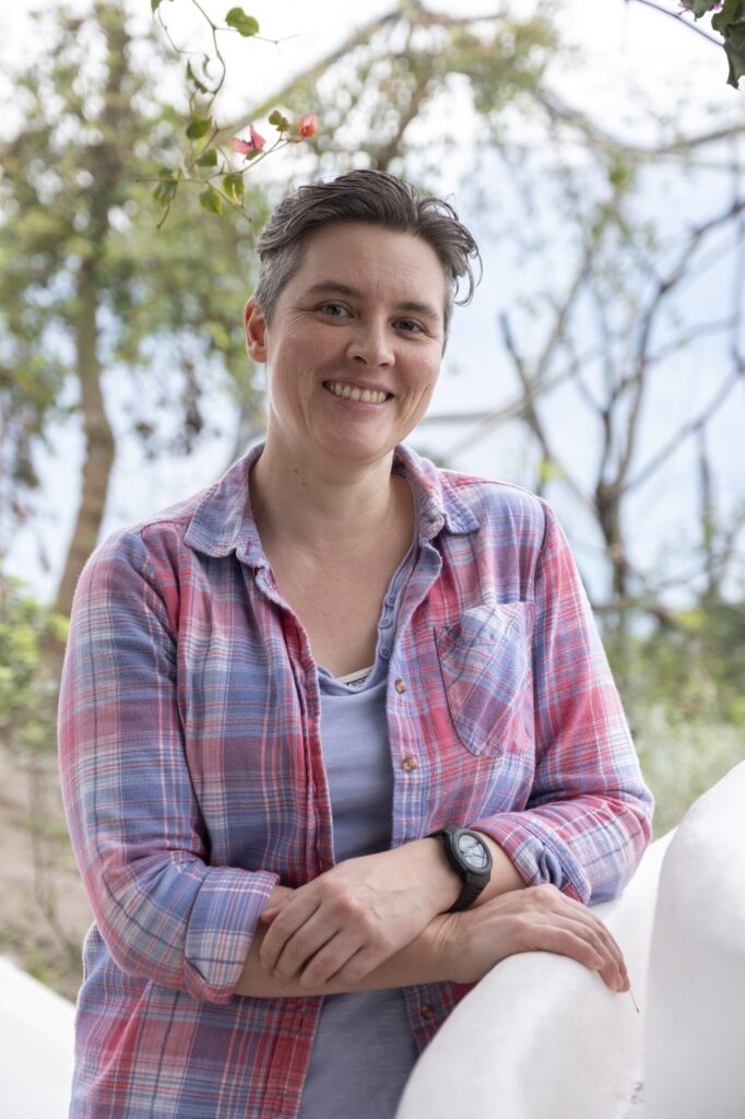 Jaiye wearing a pinkish blue checked shirt while leaning against a white wall at the Eden Project